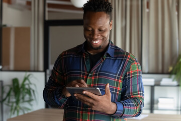 Obraz premium Smiling young African American businessman use tablet text message online on gadget. Happy millennial mixed race male employee hold pad device communicate with client on internet. Technology concept.