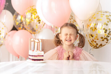 Obraz na płótnie Canvas Make a wish.Child girl in festive dress closed eyes with strong emotions making birthday wish before blowing out candles on birthday cake. Balloons background.Child's birthday