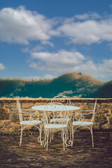 Small table with white chairs overlooking the terrace of an ancient village. Valley with mountains and sky with clouds.