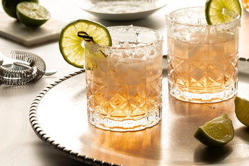 Margarita cocktails with salted rims on a metal tray with ingredients and strainer in behind.