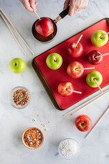 Top down view of hands dipping an apple into caramel sauce surrounded by ingredients to make...