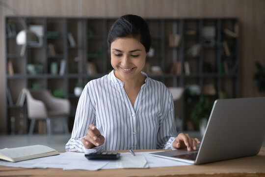Happy young Indian woman sit at desk manage household family budget on machine pay bills on laptop online. Smiling ethnic female consider count expenses expenditures. Finances, banking concept.