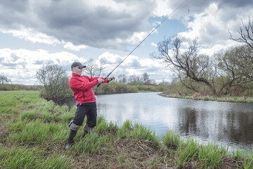 Fisherman in a red jacket is fishing on the green bank of the river.