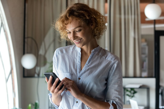 Smiling Young Caucasian Woman Look At Cellphone Screen Browse Wireless Internet On Modern Gadget. Happy Businesswoman Or Employee Hold Smartphone Talk Speak On Digital Video Call On Device.