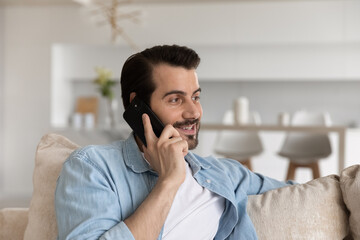 Happy positive man engaged in conversation on cellphone. Millennial guy relaxing on couch at home making telephone call on mobile phone, talking, smiling. Client consulting on helpline