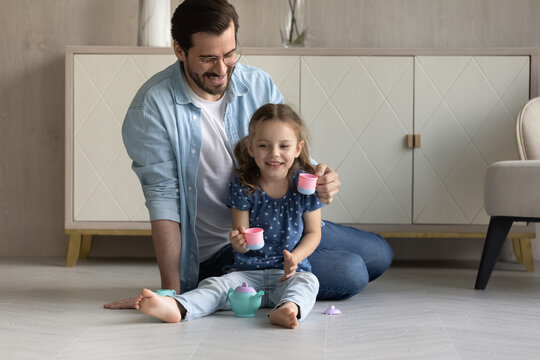 Happy dad and cute daughter girl drinking imaginary coffee from toy plastic cups, playing tea party at home, enjoying game activities together on heating warm floor. Parenthood, entertainment