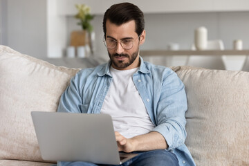 Serious young millennial man in glasses using laptop, relaxing on couch. Remote employee, self employed professional working at computer from home, chatting online, watching training webinar