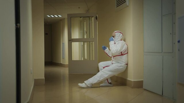 Exhausted doctor sits down on the floor in the end of working day, hospital corridor. White suit and protective mask from coronavirus, slow motion, pan shot left to right
