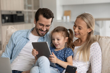 Happy excited gen Z little kid with tablet and parents with gadgets relaxing on couch, playing...