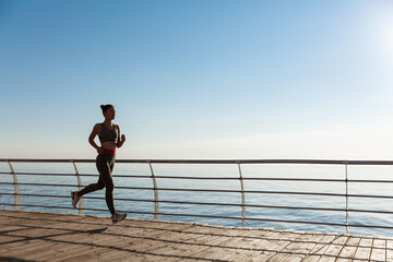 Side view of young fitness woman running along pier. Female athlete workout and jogging on the...