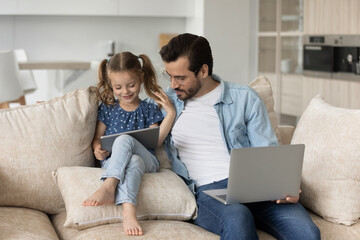 Happy gen Z little kid and daddy using tablet and laptop, sitting together on sofa at home, talking, watching media content. Dad watching daughter using app on internet. Family with gadgets