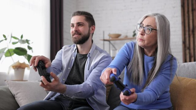 Adult son and senior mother enjoy spending time together. Grandmother and grandson playing video games using gamepads at home. Mothers Day. Two generation family. Active modern elderly people.