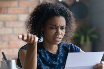 Frustrated millennial young Black woman reading paper letter with bad news. Mixed raced student...