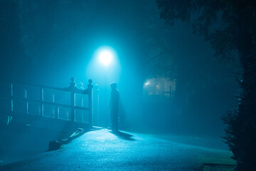 A spooky hooded figure, silhouetted against lights on a path at night. With a moody edit.