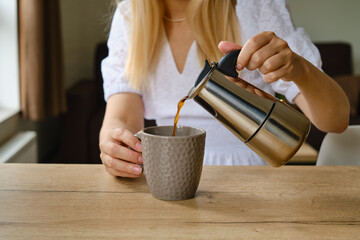 Girl pours coffee from a stylish moka pot into a cup. Equipment for making coffee. Morning coffee for breakfast. Fragrant black coffee in a cup close up