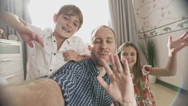 Selfie portrait of family of three smiling and laughing to camera. Wide angle view shot of father, son and daughter video chats on smart phone on living room background.