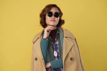 Portrait of stylish fashionable young woman in warm coat and sunglasses standing against yellow background