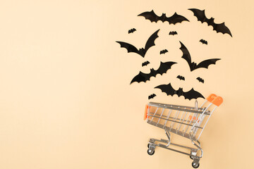 Top view photo of shopping cart model with flying bats silhouettes on isolated beige background with copyspace