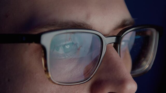 Closeup of man broker wearing eyeglasses in front of the screen analysing stock market changes, concentrated look. Young man busy working alone at night, gimbal shot