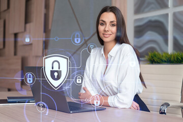 Attractive businesswoman in white shirt at workplace working with laptop to defend customer cyber security. Concept of clients information protection. Padlock hologram over office background.
