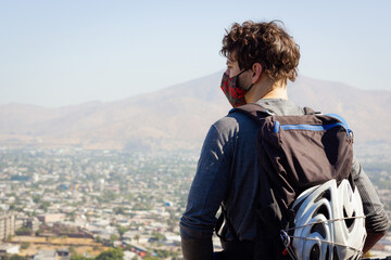 Young biker with face covering and helmet on backpack looking at city views from top on sunny day. Sports man taking a rest while enjoying landscape