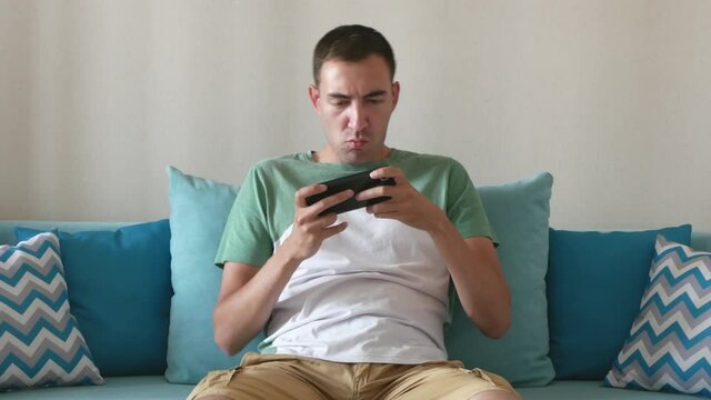 Young man plays on smartphone and wins. Concentrated man plays on smartphone and rejoices of win while sits in sofa at home.