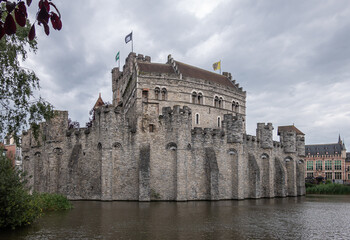 Fototapeta na wymiar Gent, Flanders, Belgium - July 30, 2021: Gray stone Gravensteen medieval castle under gray cloudscape behind its dark water moat. Some green foliage and colored flags.