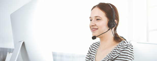 Casual dressed young woman using headset and computer while talking with customers online in sunny...