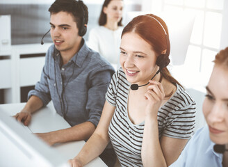 Casual dressed young woman using headset and computer while talking with customers online. Group of operators at work in sunny office. Call center