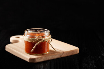 Authentic Spanish Sofrito sauce made with tomato, peppers, onion, garlic, herbs and olive oil in glass jar on cuttery board. Dark wooden background, side view