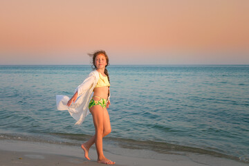 A young girl walks along the sea coast at sunset. A child in a long white shirt enjoys the sea breeze.