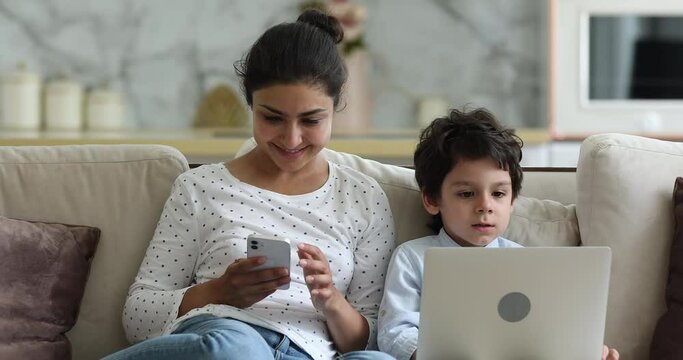 Indian woman her little son sit on sofa use gadgets. Mom hold phone 4s boy put laptop on laps enjoy online fun play video games. Devices overuse, young generation internet technology addiction concept