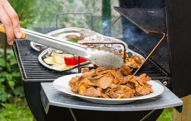 Grilling of hot pork meat and vegetable slices on charcoal grill on garden bbq party. Detail of human hand holding metal tongs and delicious barbecue meal on plate or alu trays on steel cooking grate.