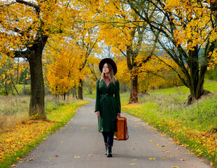 Fototapeta premium Woman in green cloak with suitcase on countryside road in autumn season time