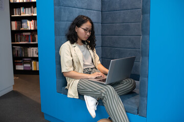 Young pretty student networking with laptop on her knees in contemporary bookstore