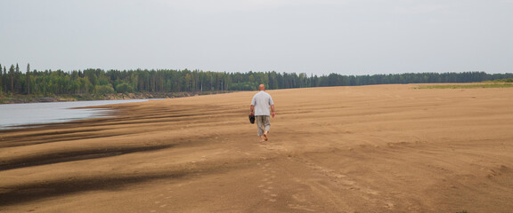 A man walks along a sandy beach along the river bank.