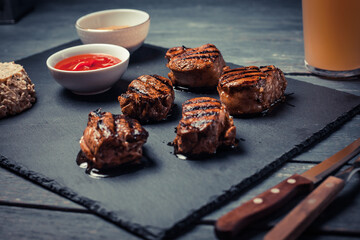 Grilled pork medallions with cheesy salad, ketchup and mustard on dark stone board. Served with glass of beer.