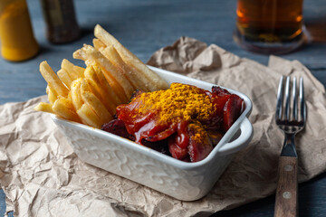 Currywurst with french fries in white bowl on craft paper served with beer on wooden table close up.