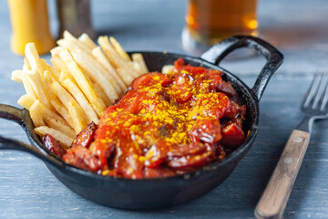 Currywurst with french fries in black pan served with beer on wooden table.