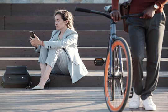 Serious businesswoman in formalwear texting in smartphone with young man pushing bicycle in front