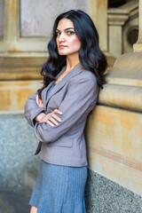 Dressing in a gray knit under dress and a jacket, crossing arms, a young businesswoman is standing outside an office building, looking sideways, relaxing and thinking.