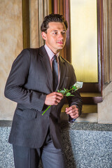 Standing by a window and dressing formally , a young handsome guy is holding a white flower and...