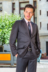 Dressing formally, two hands putting in pockets,  a young businessman is standing outside an office building, confidently looking forward..