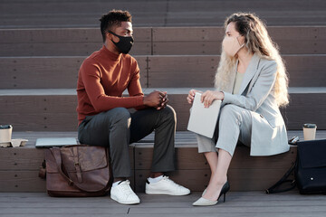 Two young contemporary business people in protective masks interacting at outdoor meeting