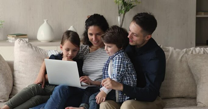 Happy bonding two different generations family using computer, resting on couch at home. Sincere affectionate young couple parents watching cartoons, shopping online or web surfing with children.