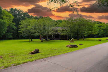 a brown log cabin in the park near a paved road surrounded by lush green grass and trees with powerful clouds at McIntosh Reserve Park in Whitesburg Georgia