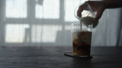 pour milk over cold brew coffee over ice cube in tumbler glass on wood table