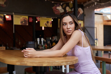 beautiful young asian woman with dark long hair in light pink dress sitting at table of dinery, having rest. pretty student lady having cup of coffee at cafe veranda. summer chill, rest, lifestyle