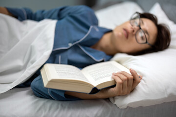 Middle aged woman in eyeglasses and pajams enjoying sweet nap on comfy bed with book in hands. Caucasin brunette fall asleep while reading at night.