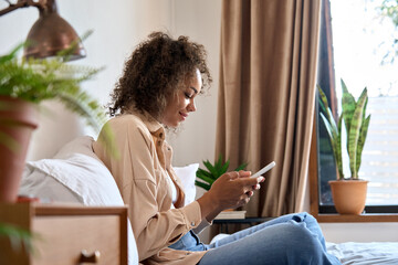 Young happy African American woman using cell phone sitting on bed. Smiling mixed race girl holding smartphone online mobile applications surfing social media app, texting, doing e shopping in bedroom